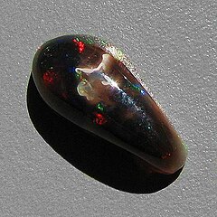 Een cabochon in vrije vorm van zwart Virgin Valley-houtvervangend opaal met rood, blauw en groen vuur dat afsteekt tegen het donkere basisopaal.