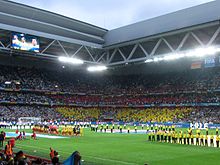 Photographie en couleur d'un angle du stade avec, à gauche, les équipes d'Allemagne et d'Ukraine, et au fond, la foule aux couleurs de l'Ukraine dans les gradins