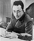 Albert Camus Albert Camus, gagnant de prix Nobel, portrait en buste, pose au bureau, faisant face a gauche, cigarette de tabagisme.jpg