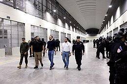 Nayib Bukele surrounded by four government officials while touring through a prison cell block, with prison guards in riot gear are standing in formation in front of cell entrances