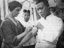 A white woman administering a vaccination to the arm of a Chinese man, while a young Chinese person looks on from the background