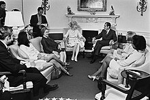 Five women and President Richard Nixon seated in an informal circle in front of a fireplace