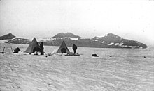 Photo en noir et blanc de deux tentes pyramidales sur un glacier devant un massif montagneux peu élevé.