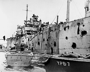 Salvage and rescue work underway on Mindanao shortly after Mount Hood blew up about 350 yd (320 m) away. Note the heavy damage to Mindanao's hull and superstructure, including large holes from fragment impacts. View looks forward from alongside her port quarter. Mindanao had 180 crewmen killed and injured by this explosion. She was under repair until 21 December 1944. Small craft alongside or nearby include (from left) YPB-6 (probable identification), two LCVPs and YPB-7. USS Mindanao (ARG-3) damaged by explosion of USS Mount Hood (AE-11) in Seeadler Harbor on 10 November 1944.jpg