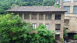 Vue de face de l'usine d'Entraygues sur trois niveaux avec ses sept grandes fenêtres carrées ou rectangulaires par étage et son toit en tuiles sombres, beaux arbres au premier plan