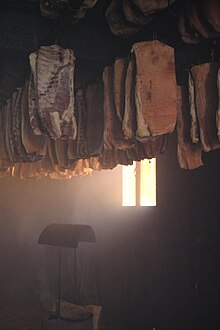 Im Dämmerlicht eines Raumes, der von einem einzigen Fenster beleuchtet wird, werden Fleischstücke aufgehängt, die sorgfältig in drei Reihen ausgerichtet sind.