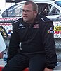 A stoutly-built man with a crew cut in a firesuit and glasses sits in front of a chain link fence, with a race car behind him.
