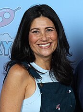 A dark-haired white woman wearing a white tank-top and blue overalls smiling towards the camera.