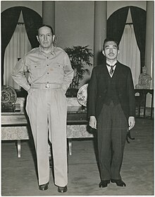 A tall Caucasian male (MacArthur), without hat and wearing open-necked shirt and trousers, standing beside a much shorter Asian man (Hirohito) in a dark suit.