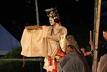 Acteur debout en tenue traditionnelle rose et masque blanc, un second assis de dos au premier plan.