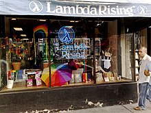 Storefront with an awning reading "Lambda Rising" with "Lambda Rising" neon sign in the window, surrounded by LGBT-themed products.