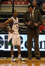Um homem, vestindo um terno marrom, está conversando com um jogador de basquete na lateral de uma quadra de basquete.