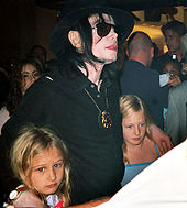 A man wearing a black fedora and dark aviator sunglasses places his hands on the shoulders of two young girls. His black hair and shirt contrast with his pigment-less skin. The man sports a pendant around his neck, as well as a watch on his right wrist. He is surrounded by a crowd of people.