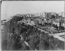 A black and white photograph of Riverside Drive, New York. Train tracks can be seen on the left of a photograph, with a strip of trees (perhaps a park). On the right side of the photograph are large buildings stretching out of frame and into the distance.