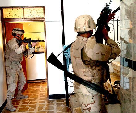 US soldiers in Tal Afar, Iraq, search for insurgents. The soldier in the foreground is carrying an assault rifle and a shotgun on a sling for breaching
