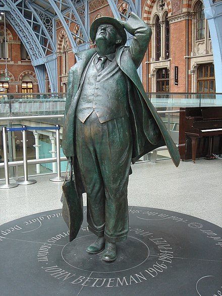 A large, bronze statue of a man. He is dressed in a suit, coat and hat and is looking up at the roof of a building.