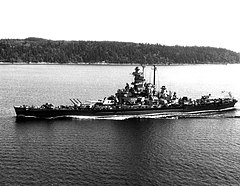 A battleship sails on calm waters with land in the background.