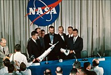 Sept hommes posant sur une estrade aux couleurs de la NASA et tenant des maquettes d'engins spatiaux.