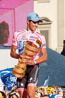 A smiling man of about thirty wearing a pink cycling jersey and a pale blue baseball cap, holding a large golden trophy.