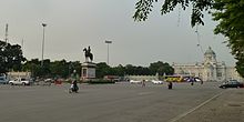 A large plaza with a bronze statue of a man on a horse in the centre; beyond the plaza is a large two-storey building with a domed roof, arched windows and columns