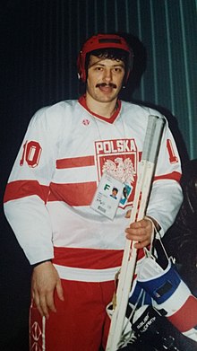 Ein männlicher Eishockeyspieler posiert für ein Foto, das von den Knien aus sichtbar ist. Er ist in Eishockeyausrüstung gekleidet und trägt das polnische Nationaltrikot. Während sein Helm aufgesetzt ist, hält er seine Handschuhe.