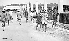 Photographie en noir et blanc de personnes sur une route.  Trois hommes sur le côté droit de la photo portent des fusils.