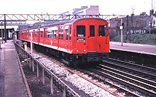 Et tre fjerdedels fotografi av et rødt O-lager med skyvedører og flared sider, og forlater Barking-stasjonen på vei til Ealing i 1980