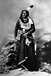 Photographie en noir et blanc d'un Amérindien en habit traditionnel coiffé d'une plume et tenant un tomahawk.