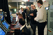Men sitting at computer terminals with other men and a woman standing behind them