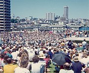 A large crowd has gathered in an open space in an urban setting (tall buildings are visible). Far away can be seen a platform, and a banner reading SHAME FRASER SHAME.