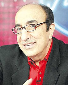 Elias Rahbani wearing glasses, red button-down shirt, and black blazer, smiling at camera with head tilted and microphone pinned to shirt.