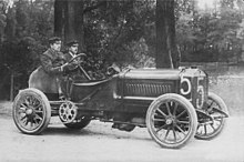 An open-topped sports car, with the number 5 written on the front and side. A driver sits on the right side of the cockpit, and his riding mechanic sits alongside.