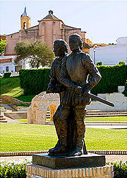Estatua de los Hermanos Pinzón en Palos de la Frontera; al fondo se observa la Fontanilla y la Iglesia de San Jorge Mártir.