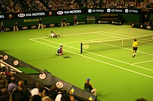 Two tennis players on a green hard court, one lying on the ground, the other standing next to the net, surrounded by crowded bleachers