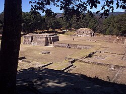 View across a series of neatly maintained low ruins, consisting of a labyrinthine series of overlapping rectangular basal platforms. Two small pyramid structures dominate the view, with pine forest providing the backdrop.