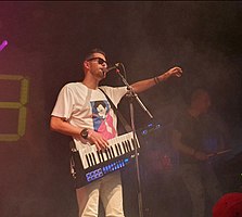 Michael Oakley wearing a white print t-shirt, light-coloured pants, and dark sunglasses, standing onstage with a keyboard around his neck, speaking or singing into a microphone, with one arm held out towards the audience