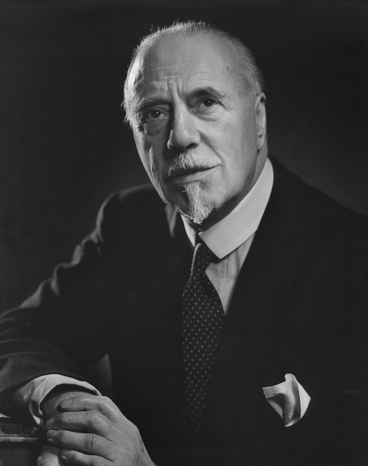 elderly white man with white receding hair and very small moustache and imperial beard， in contemporary lounge suit， facing the camera but not looking directly at it