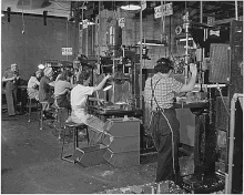 A World War II-era photo of five women working at drill presses, all wearing pants, work shirts, hats, and aprons