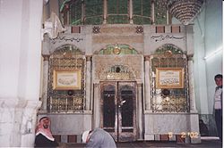 O interior de um edifício religioso mostrando dois homens sentados em frente a um recinto de mármore fechado coberto de vidro esverdeado