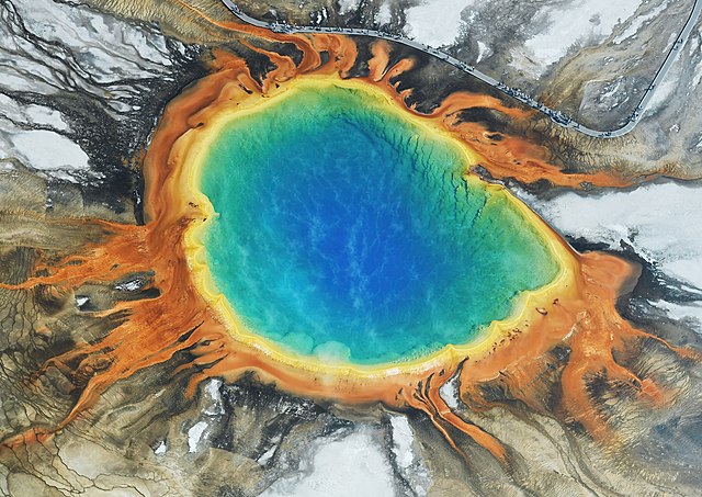 un lac dans un cratère dont les eaux montrent un gradient de couleurs du bleu foncé au centre au vert, jaune puis orange sur les bords.