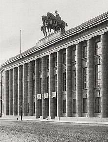 Seitliche Schwarzweißfotografie eines Gebäudes mit Säulenfassade. Zwei Pferdefiguren und zwei Männerfiguren stehen auf dem Dach.