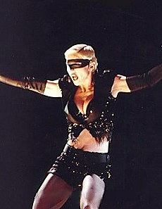 Madonna wearing black shorts and looking to her right. Her arms are open and a headset microphone to her mouth.[20]