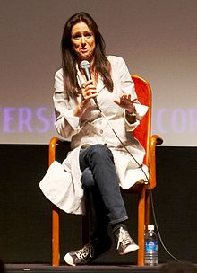 Taymor in white overcoat, jeans and sneakers, holding microphone, seated, facing camera