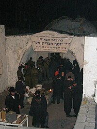 Une photographie en couleur montrant un groupe d'hommes vêtus de noir et portant des chapeaux, ainsi qu'un groupe de soldats en uniforme, debout sous une ouverture voûtée blanchie à la chaux avec une porte derrière et un dôme bas faiblement visible à l'arrière-plan