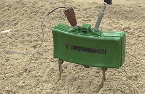 A green plastic-body mine supported by a pair of scissor legs, with "К ПРОТИВНИКУ" (K PROTIVNIKU; to the enemy) stenciled on the front.