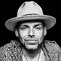 black-and-white picture of man with contour facial hair and two earrings, wearing a brimmed hat, looking at camera