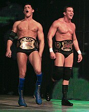 Two dark-haired men are both wearing black wrestling tights. One wears knee-high blue wrestling boots with blue knee-pads and a black elbow pad on his right arm, and the other wears black knee-high wrestling boots with black knee-pads, and white tape around his wrists. The two are wearing professional wrestling championship belts around their waists.