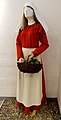 Mallinukke, jolle on puettu punainen pitkähihainen, nilkkapituinen mekko, jonka rinnassa nappi, vyötäröllä valkoinen esiliina ja päässä hiukset paljastava valkoinen huivi.