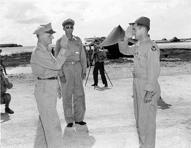 A man in a flight suit and peaked cap exchanges salutes with a man in uniform. Multiple photographers capture the moment.