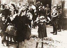 A small crowd scene in black and white. Several men, women and children are holding their hands up. The women are wearing heavy, dark coats, and carrying bags or bundles. Some of the men, women, and children have scarves or caps on their heads. Behind them, men are standing in uniforms. One is carrying a large gun. In the forefront, there is a small boy. He is wearing a cap and a buttoned-up coat, short trousers, long socks, and boots. There is a satchel over his right shoulder. His arms are in the air, with the palms of his hands facing the camera. He looks afraid. Cobblestones are visible on the ground.